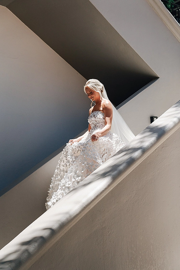 Bridal portrait of a bride on stairs in a strapless 3D floral appliqué gown with veil and earrings, in a sunlit white stairwell