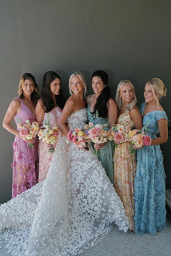 Bride with bridesmaids portrait showing bride in lace appliqué gown with long train, bridesmaids in pastel dresses holding rose bouquets by gray wall