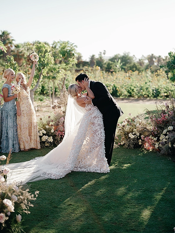 Wedding kiss as the groom dips the bride in her veil and lace dress, bouquet raised, bridesmaids cheering on a garden lawn