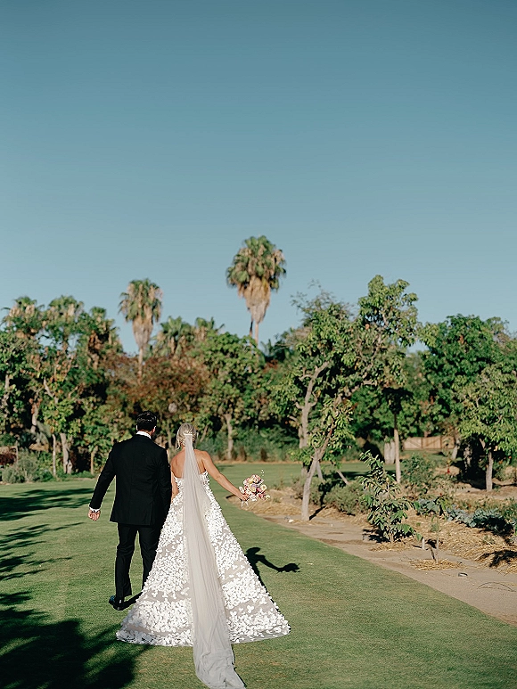 Couple portrait of bride and groom walking away hand in hand, her long veil and pastel rose bouquet trailing on a palm-lined lawn path
