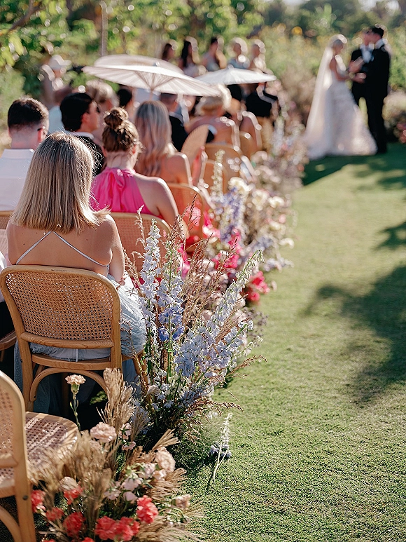 Outdoor wedding ceremony with bride and groom at the altar, aisle florals lining a sunlit garden ceremony aisle with parasols and chairs