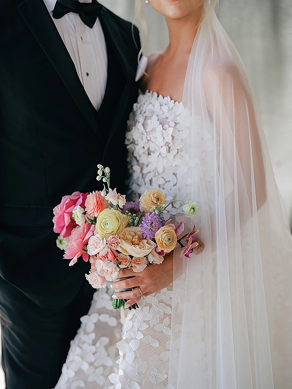 Couple portrait of bride and groom close up, her bouquet held forward with veil drape, against a soft neutral wall backdrop