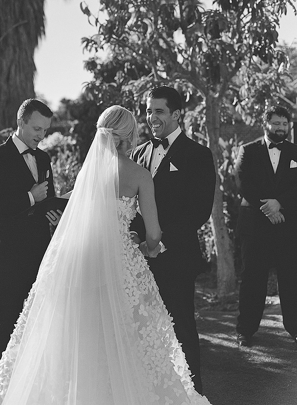 Wedding vows at an outdoor wedding ceremony as bride in strapless lace dress and long veil faces tuxedoed groom in sunlight garden