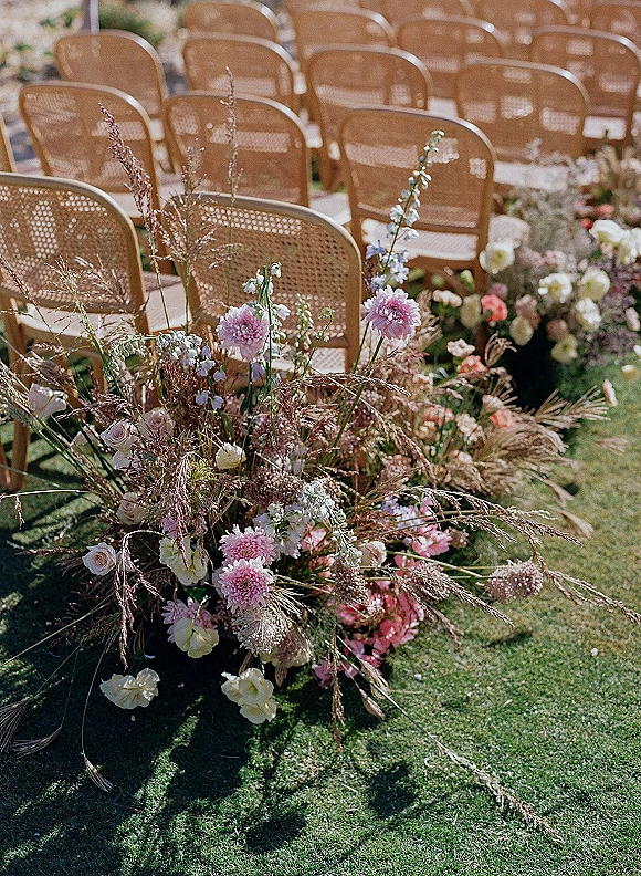 Ceremony aisle decor with outdoor ceremony aisle flowers lining wicker chairs, featuring low pink and white blooms with dried grasses on a lawn