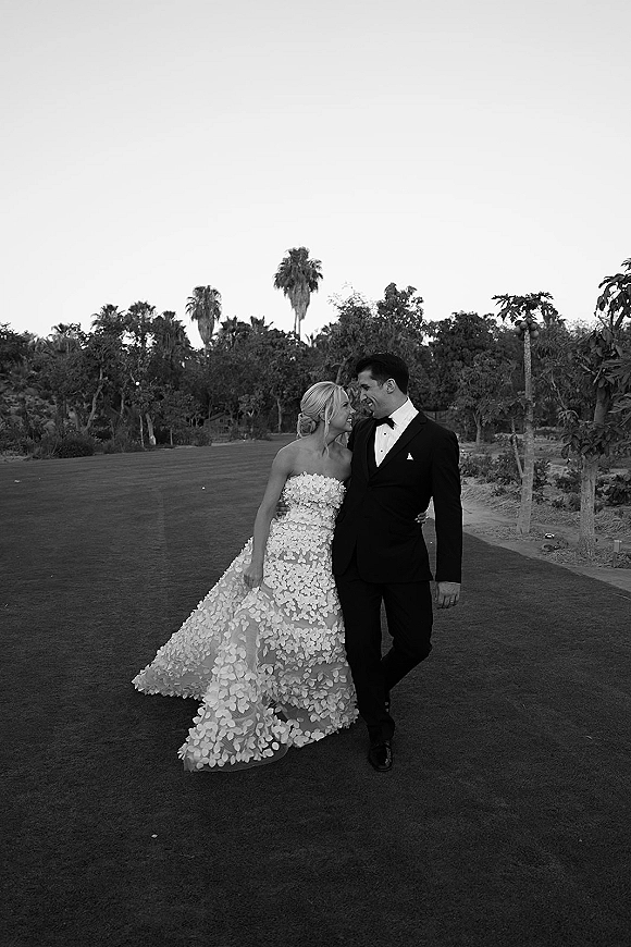 Couple portrait in black and white of bride and groom walking and laughing on a garden path with palm trees and open sky behind