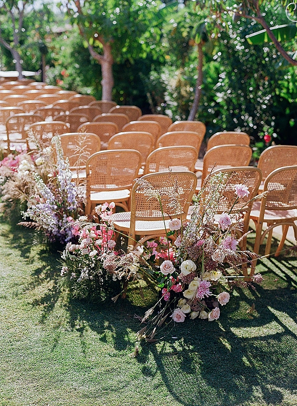 Ceremony aisle decor with ground floral aisle of roses, garden flowers, and greenery lining wood chairs on a sunlit grass lawn