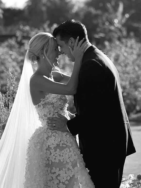 Couple portrait in a black and white wedding portrait, bride and groom touch foreheads while embracing in a sunlit garden bokeh backdrop