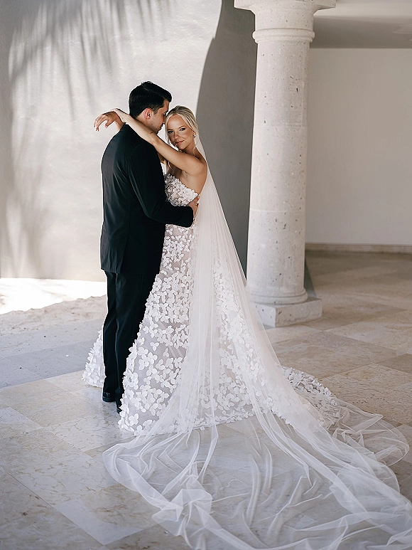 Couple portrait of bride and groom embrace, her strapless lace gown and long veil train flowing in a sunlit corridor by stone columns