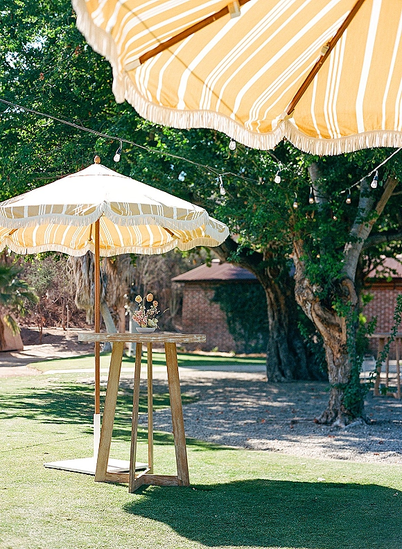 Outdoor cocktail area with striped patio umbrellas and a high top table with small floral arrangement under string lights on a green lawn
