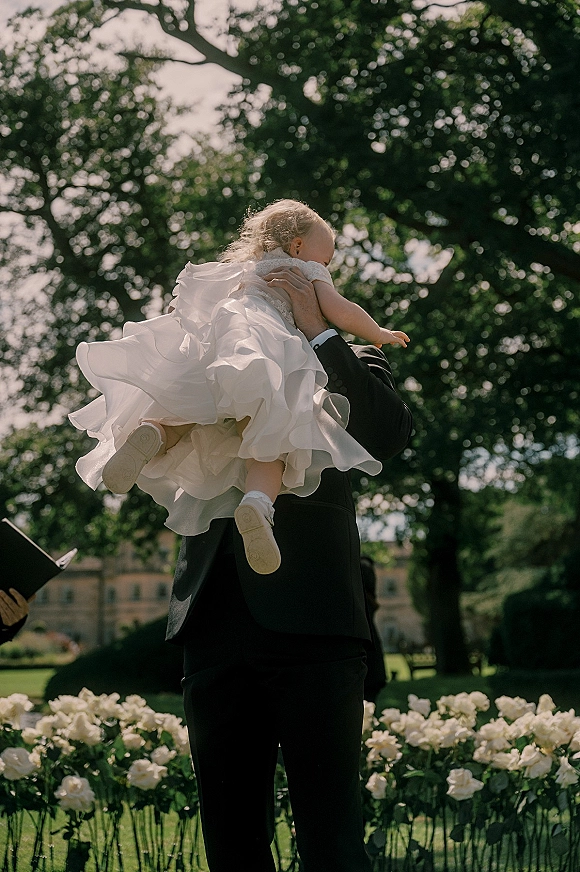Flower girl moment as groom holding flower girl in a tulle dress beside a white floral border on a sunlit garden lawn