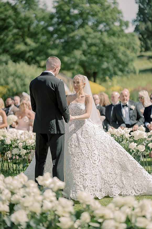 Wedding vows as bride in a strapless lace gown and long veil holds hands with groom in tux, white rose aisle florals on green lawn