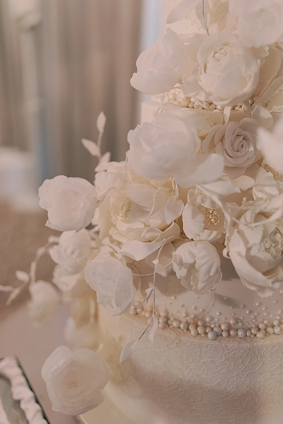 Wedding cake with textured buttercream tiers, cascading white rose sugar flowers and pearl beading on a table before soft drapery
