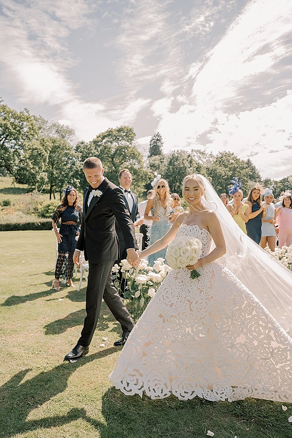 Wedding recessional as bride and groom walking aisle hand in hand, her long veil flowing, with guests and white chairs on a sunny lawn