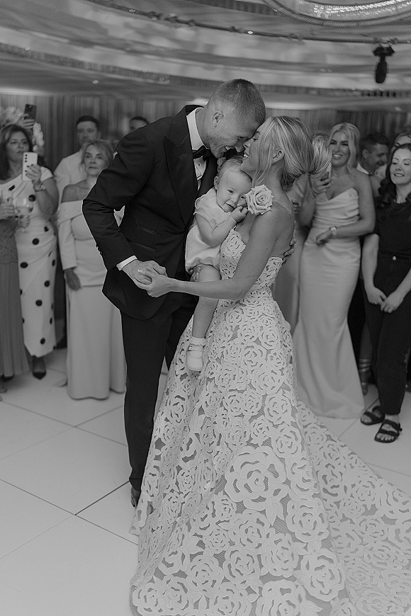 First dance moment as bride in a lace gown and groom in tuxedo sway while holding a baby, guests watching on a lit reception floor