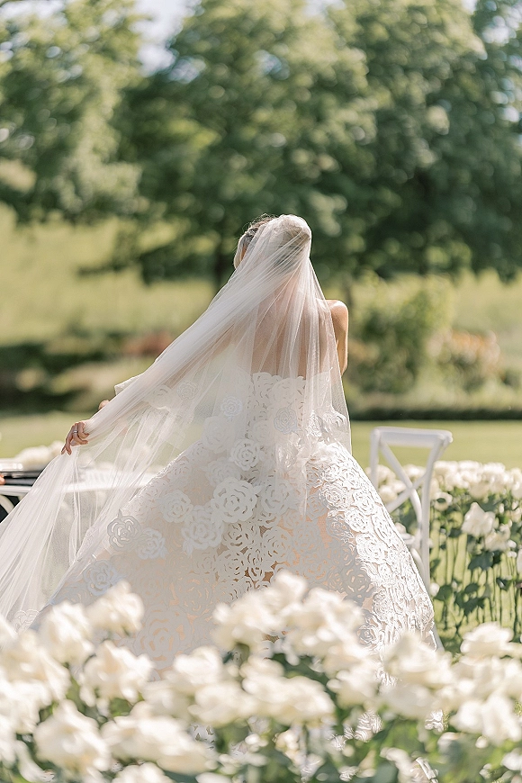 Bridal portrait of a back view bride in a lace gown holding her cathedral veil, with white chairs and trees on a green lawn