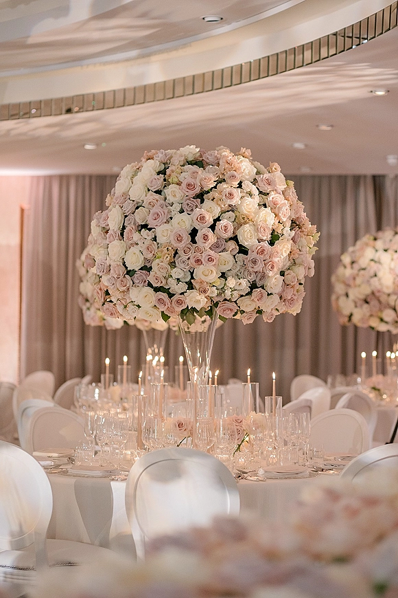 Reception tablescape with wedding table centerpiece of tall blush and white roses in clear vases, taper candles, and glassware in a draped banquet hall