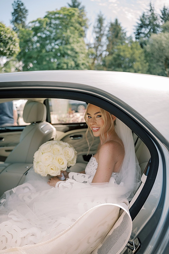 Bridal portrait of a bride in car holding a white rose bouquet in a strapless wedding dress, with veil and greenery outside the window