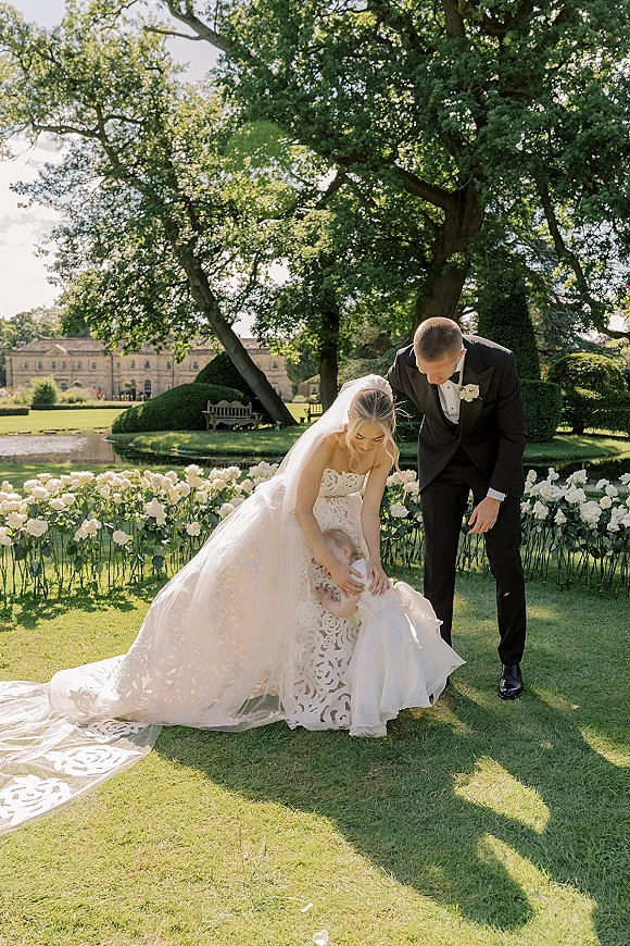 Family wedding portrait of bride kneeling in a strapless lace gown and veil beside groom in black tux, with flower girl on a sunlit garden lawn