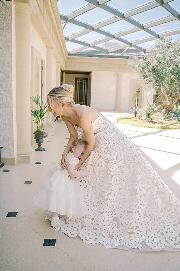 Bride with flower girl sharing a flower girl hug, showing a lace train under a glass-canopy courtyard walkway with columns and plants