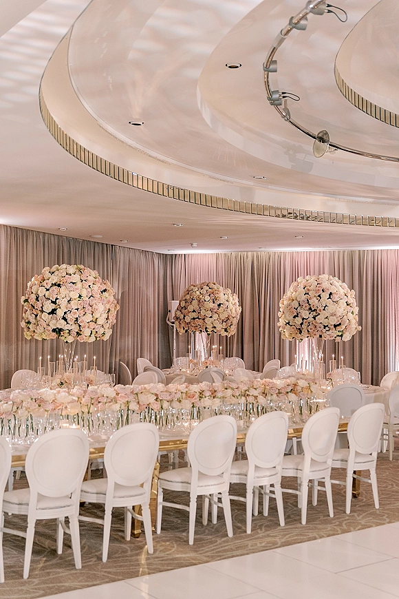 Reception tablescape with long head table, tall blush rose centerpieces in glass vases, taper candles, and gold legs in a formal ballroom