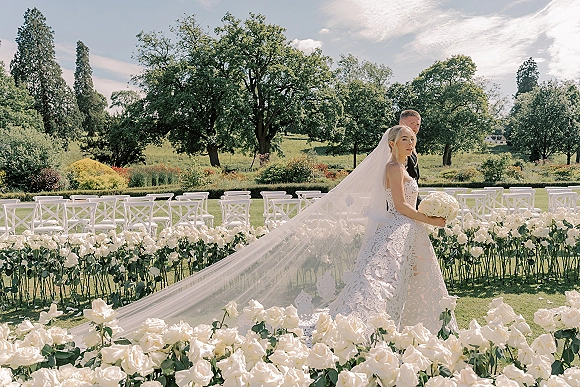 Couple portrait of bride and groom in an outdoor wedding portrait, her cathedral veil flowing as she holds a white bouquet on a garden lawn
