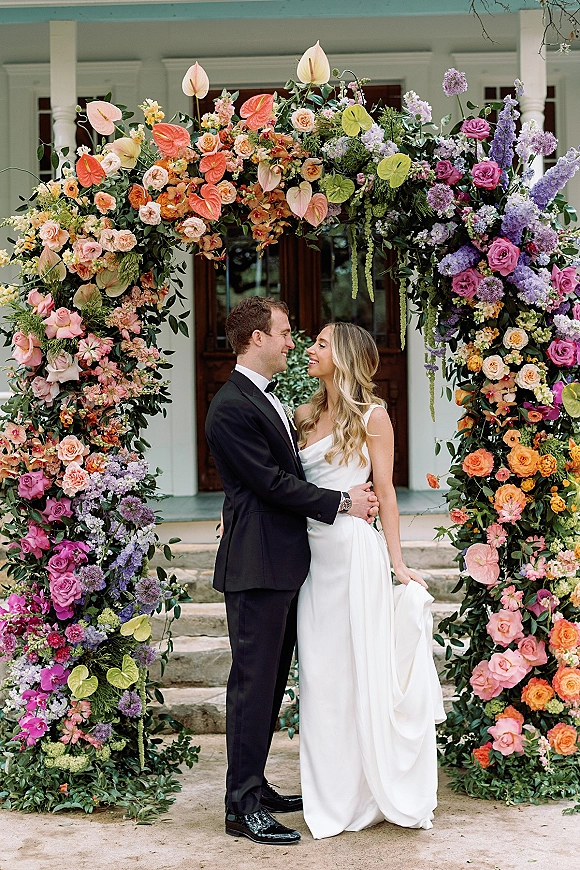 Couple portrait of bride and groom under floral arch, her strapless gown and his tuxedo framed by porch columns and stone steps