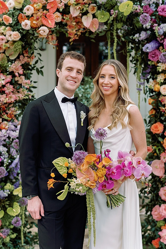 Couple portrait of bride in a white dress and groom in black tuxedo under a colorful floral arch at a greenery-framed doorway.