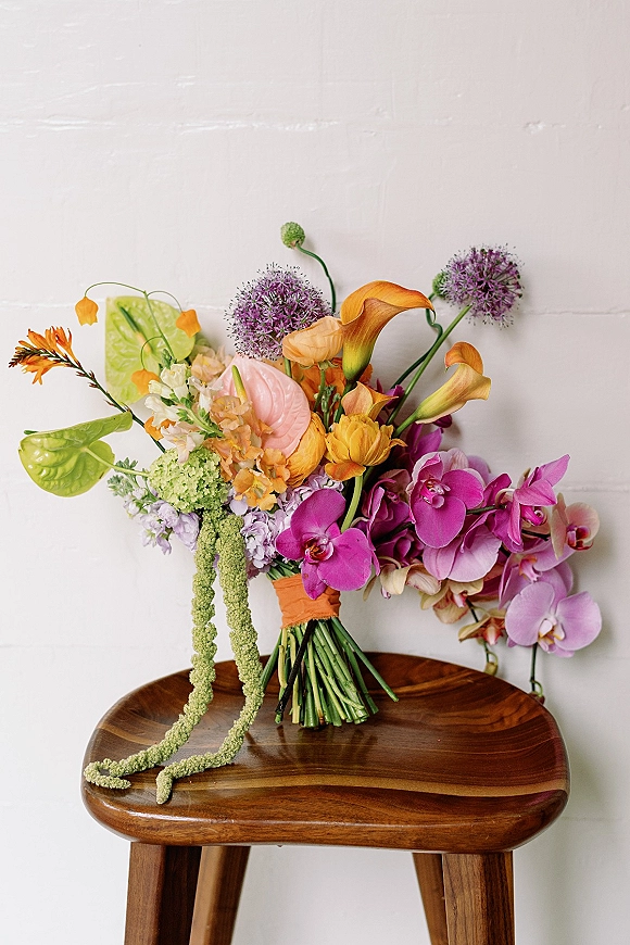 Wedding bouquet with tropical wedding bouquet blooms—orchids, calla lilies, anthurium, and trailing amaranthus—on a wooden stool by a white wall