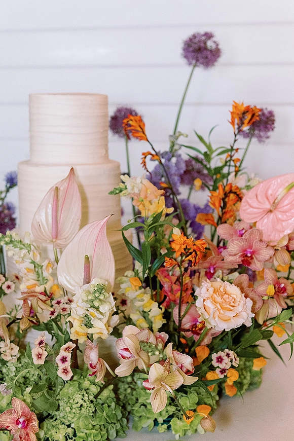 Wedding cake with textured buttercream, tiered white design topped with orchids and lush florals on a tabletop against a white wall