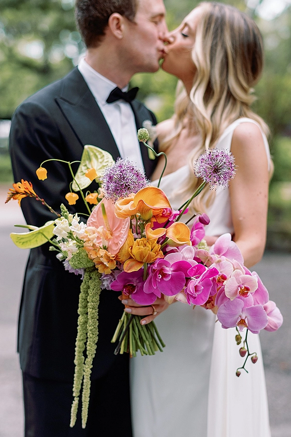 Wedding kiss portrait of bride and groom kissing, bride holding a tropical bouquet with orchids and calla lilies on a tree-lined walkway