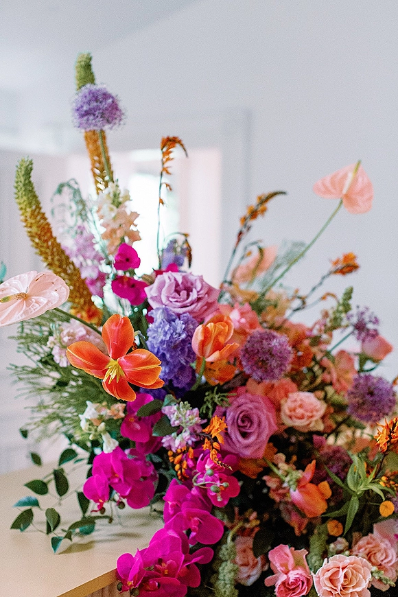 Wedding floral arrangement with colorful roses, orchids, and anthurium on a tabletop against a white wall indoors