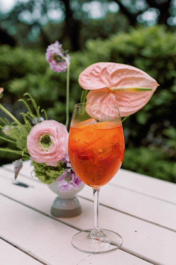 Wedding signature cocktail aperol spritz wedding served in a stemmed glass with ice beside anthurium and ranunculus centerpiece on a garden table