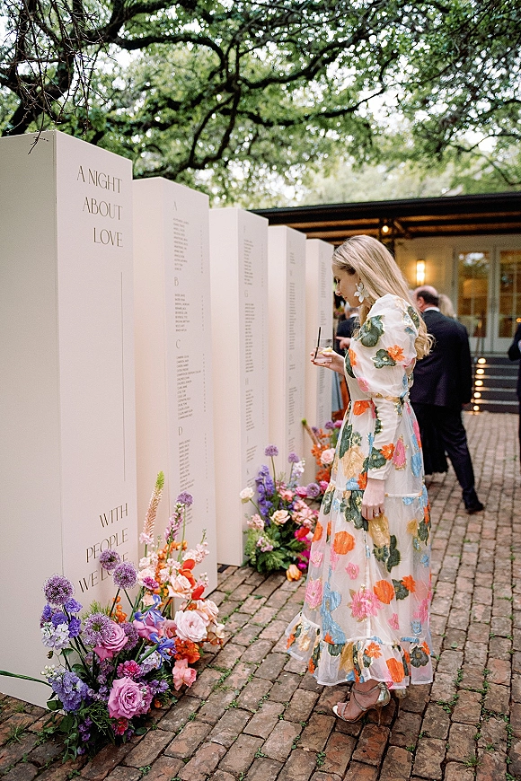 Wedding seating chart display on large panels with wildflower blooms as a guest in a floral dress checks names on a brick walkway patio
