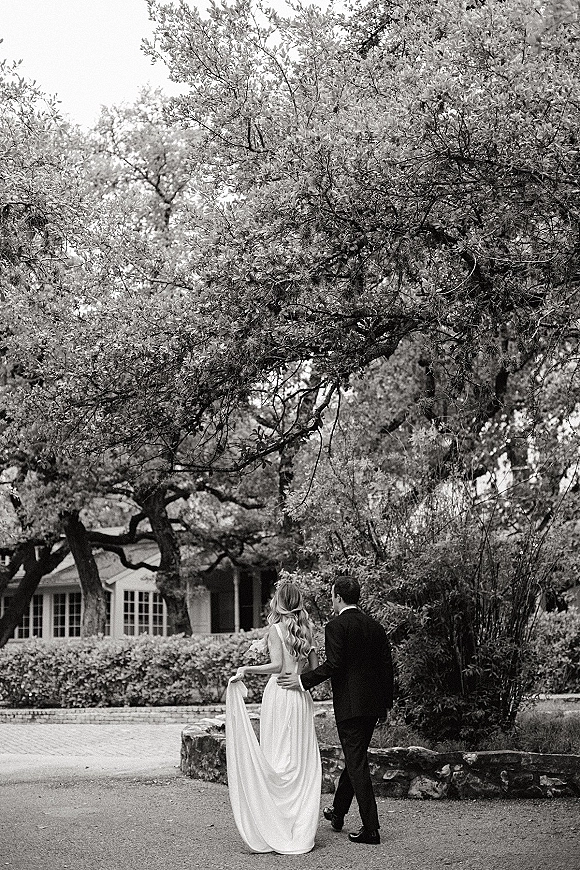 Couple portrait of bride and groom walking away, groom’s arm around her as she lifts her long dress train on a gravel driveway by a stone wall