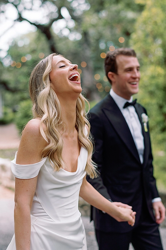 Wedding couple portrait of a laughing bride and groom holding hands, her off-shoulder dress and his tuxedo, on a garden pathway with string lights