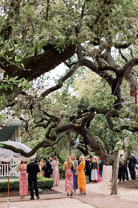 Wedding cocktail hour with outdoor cocktail hour guests mingling under string lights on a brick patio beside oak trees and porch seating