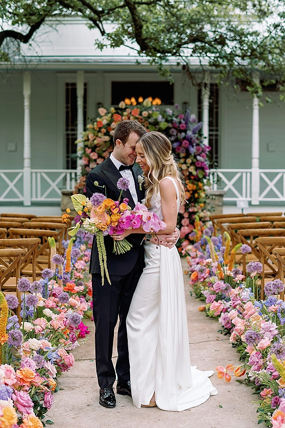 Couple portrait of bride and groom embracing, holding a colorful bouquet beside a floral arch on an outdoor aisle lined with flowers