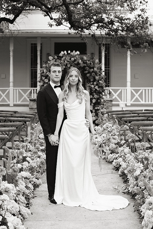 Couple portrait of bride and groom portrait holding hands on a floral-lined outdoor aisle beneath a wedding floral arch with porch columns behind