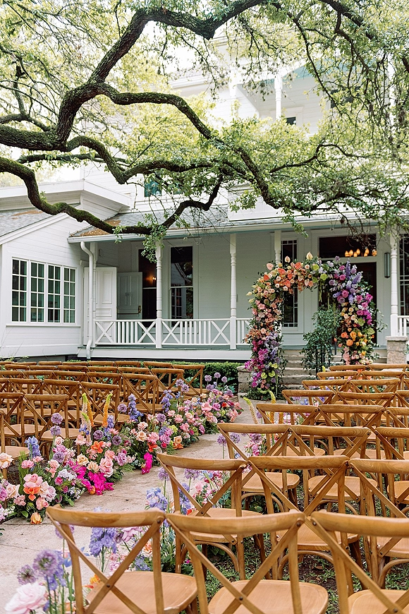 Ceremony setup with outdoor wedding ceremony crossback chairs facing a floral arch, aisle flowers and greenery on stone steps by a white porch