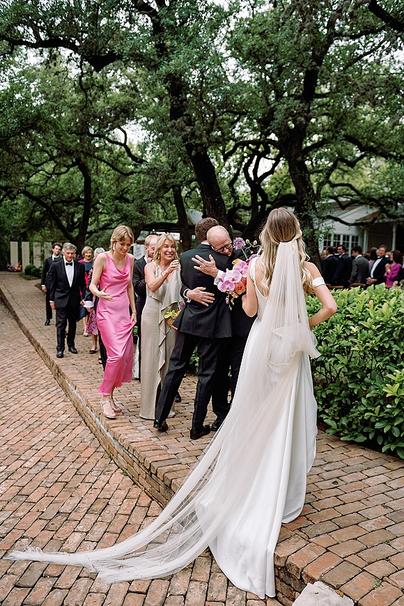 Wedding guest hugs as the bride greeting guests, holding an orchid bouquet with a cathedral veil trailing along a brick walkway courtyard