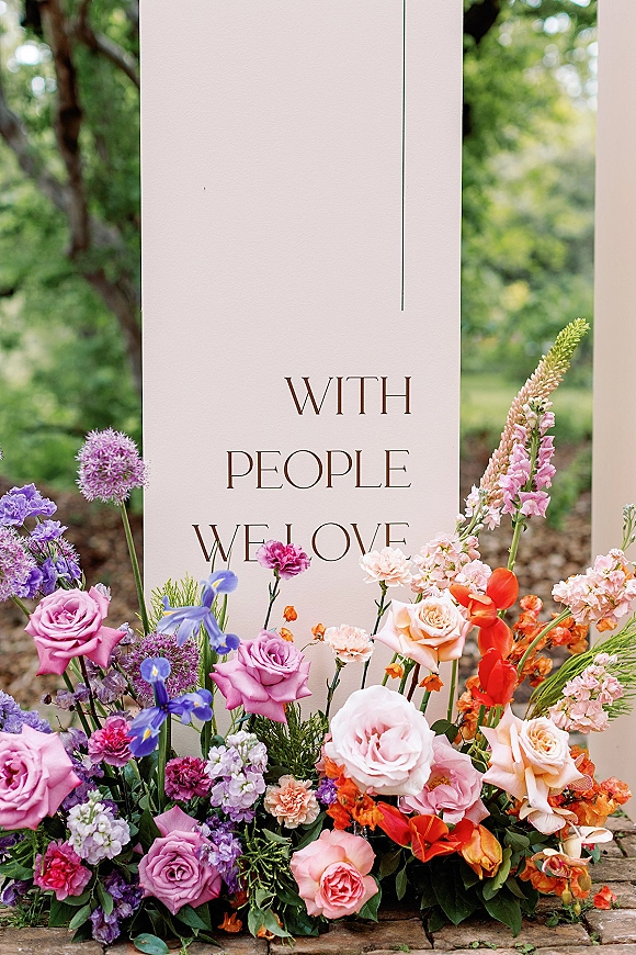 Wedding welcome sign on freestanding panels with a ground floral arrangement of roses, delphinium, and greenery by a garden path.
