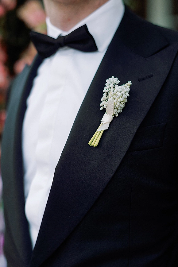 Groom boutonniere with lily of the valley boutonniere wrapped in white ribbon on a black tuxedo lapel, guests and greenery blurred behind