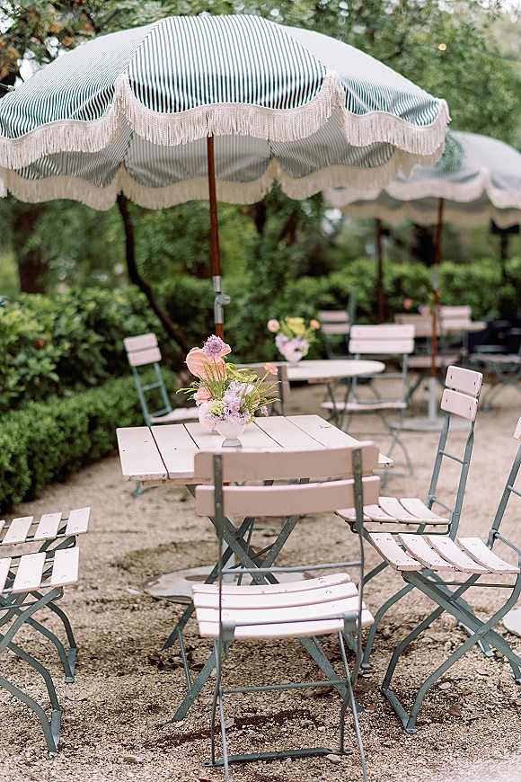 Outdoor reception seating with garden wedding bistro tables under a striped patio umbrella, folding chairs and a small floral vase on a gravel patio
