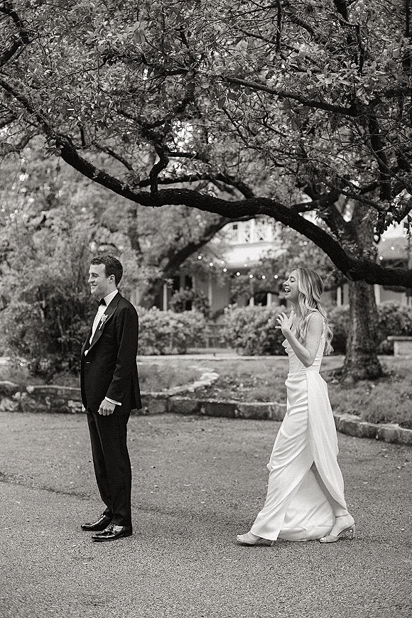 Wedding first look as bride taps groom’s shoulder, her strapless dress and heels beside his tuxedo under string lights in a garden driveway