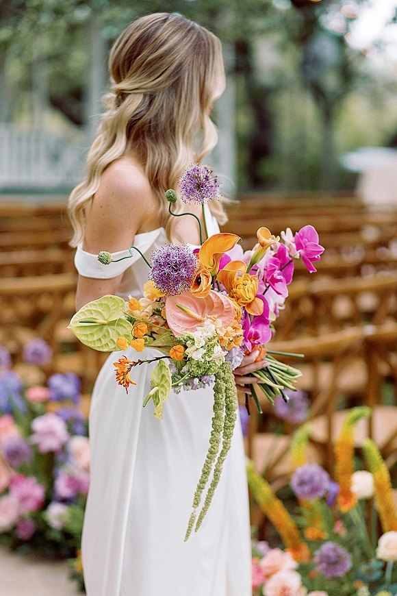 Bridal portrait of a bride holding bouquet in an off the shoulder wedding dress, with colorful orchids, by garden ceremony chairs and aisle florals