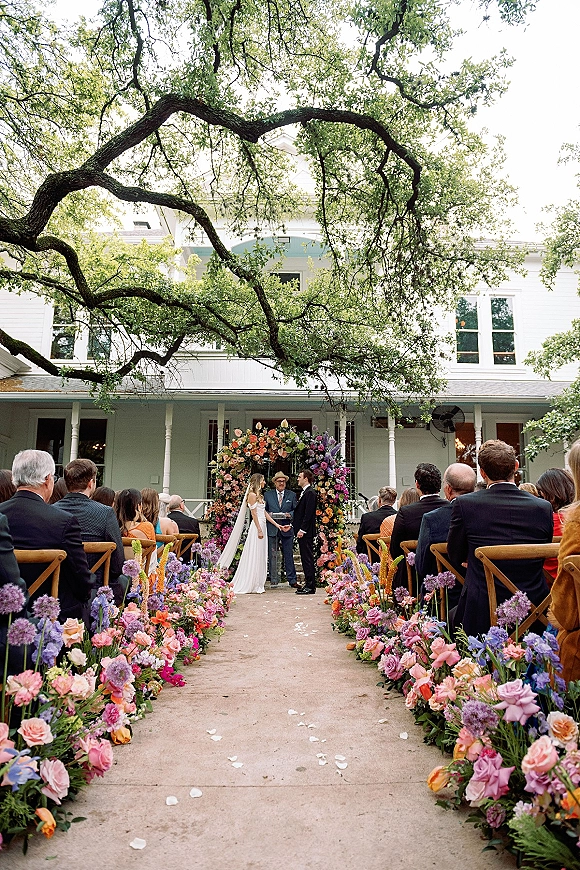 Wedding ceremony with bride and groom at the altar beneath a floral arch, with aisle flowers and rose petals in a garden setting