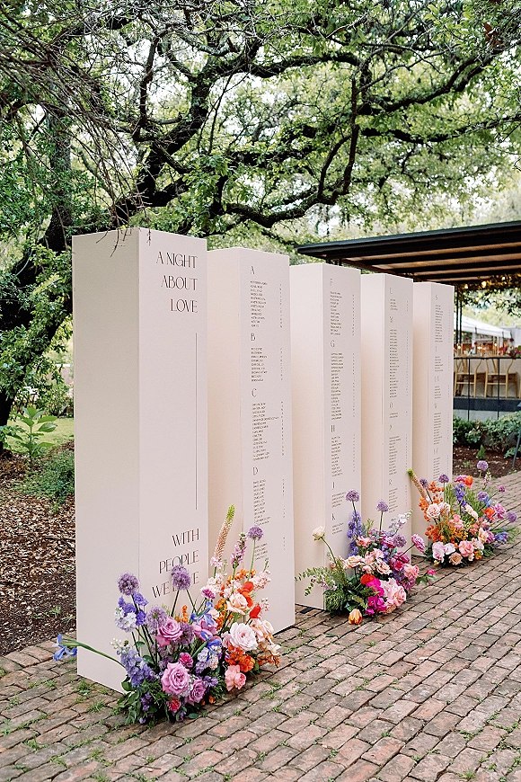 Wedding seating chart on a white panel display with printed typography and wildflower accents on a brick walkway in a garden patio setting