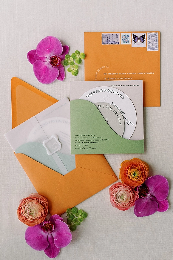 Invitation suite with colorful wedding invitations, orange and green envelopes, white wax seal, vintage stamps, orchids and ranunculus on light fabric backdrop