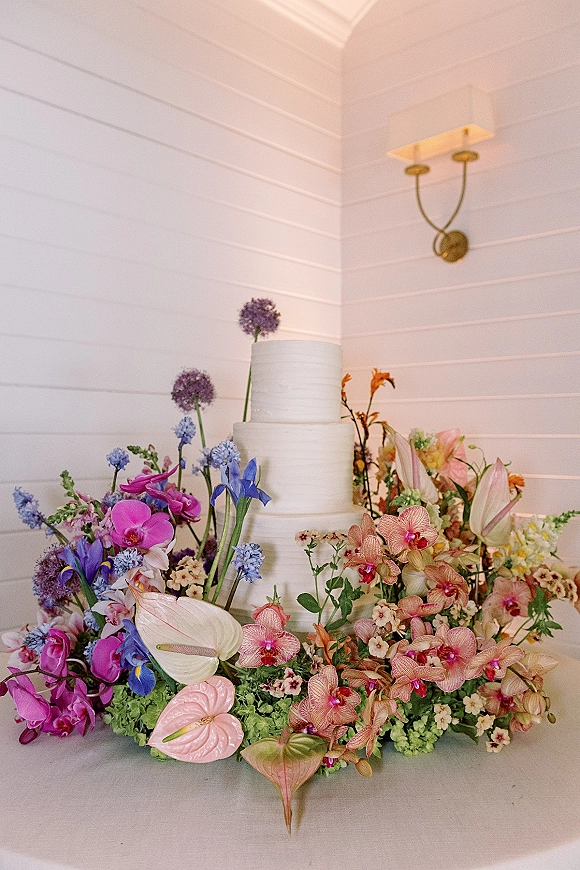 Wedding cake with orchids, a two tier white buttercream design surrounded by colorful blooms on a tabletop against white shiplap wall