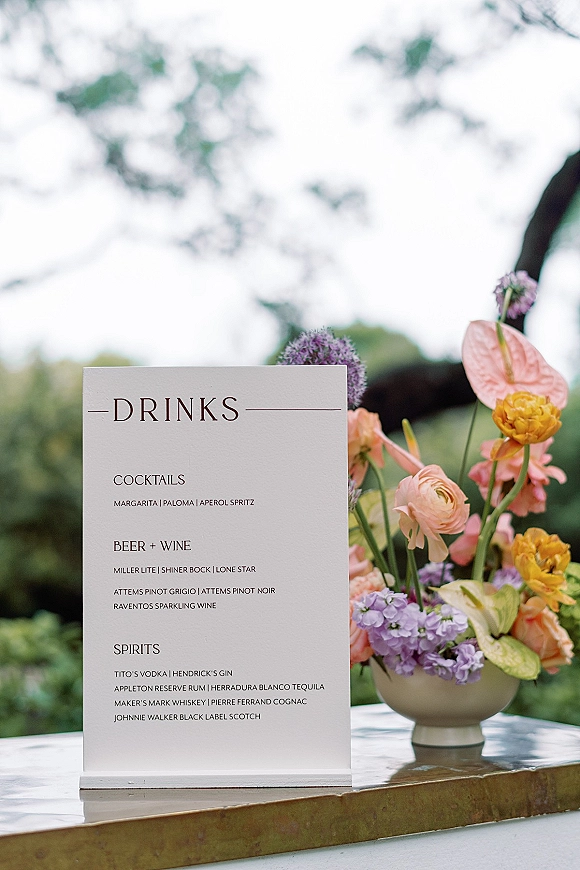 Wedding drinks menu displayed in an acrylic holder on a bar counter with a floral arrangement, set against trees and open sky outdoors